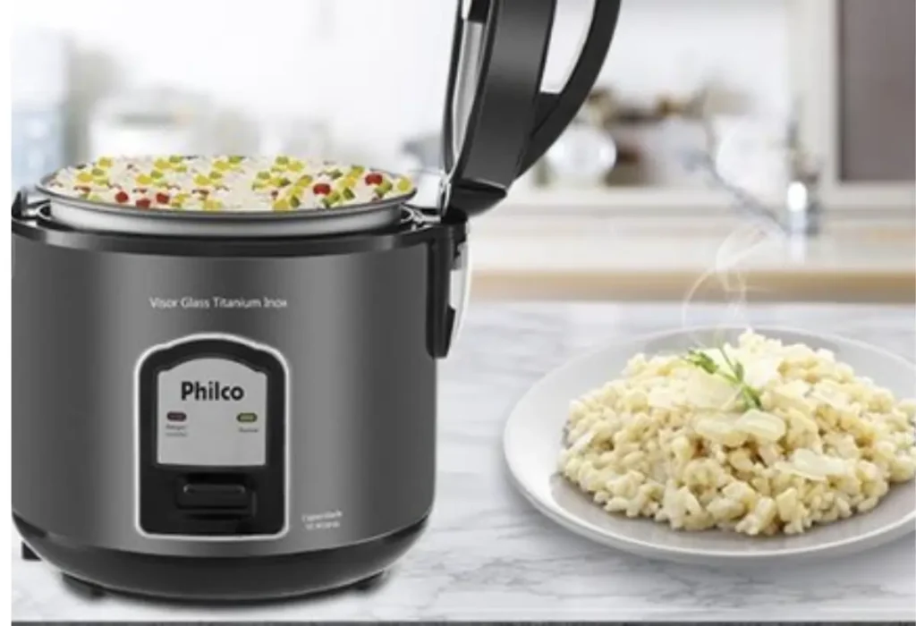 Uma panela elétrica da marca philco em uma mesa, ao lado um prato com arroz fresco.