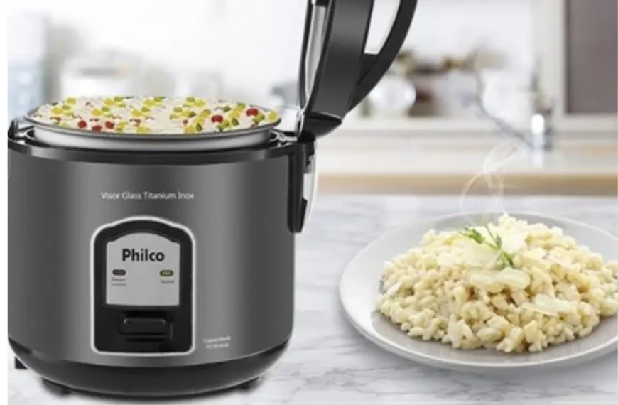 Uma panela elétrica da marca philco em uma mesa, ao lado um prato com arroz fresco.